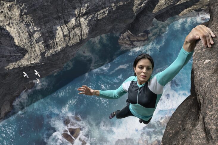 Rock climber demonstrating exceptional grip strength while hanging from cliff face above ocean water showing muscle cardiovascular connection