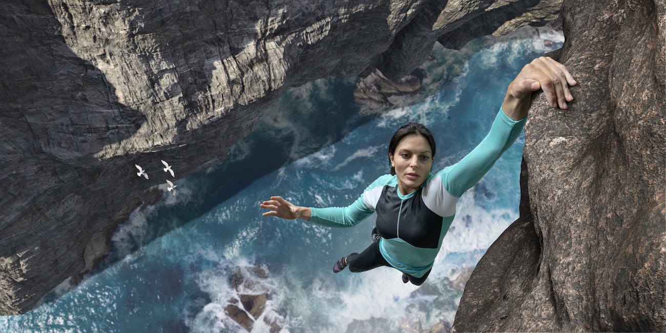 Rock climber demonstrating exceptional grip strength while hanging from cliff face above ocean water showing muscle cardiovascular connection