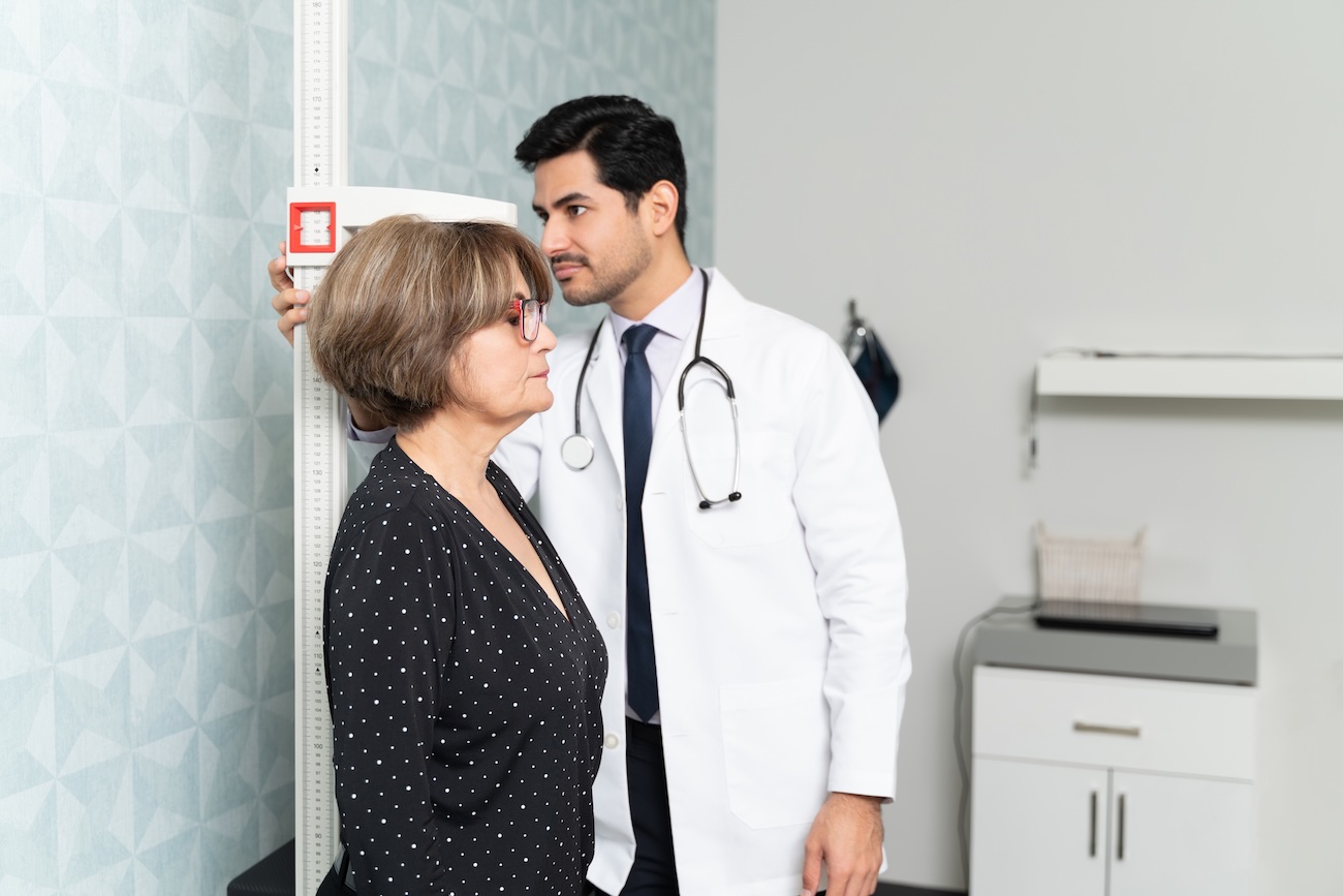 Doctor measuring elderly woman's height using wall-mounted stadiometer in medical office setting