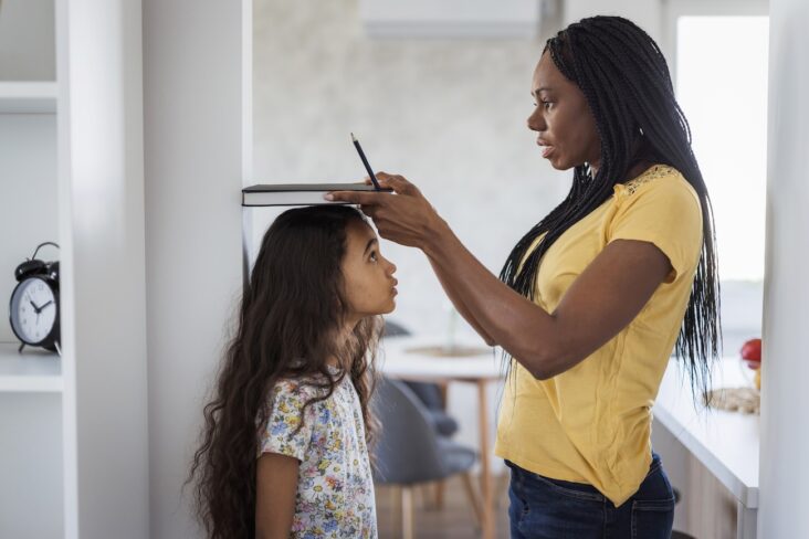 Woman measuring young girl's height at home using flat object against white wall